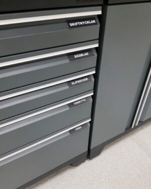 En uppsättning grå verktygslådor märkta på svenska med "Magnetiska etiketthållare" med text: "SKIFTNYCKLAR", "SÅGBLAD", "SLIPSKIVOR", "VERKTYG" och "FÄRG." Skåpet är placerat på ett ljusgrått golv.