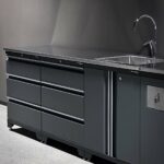 En elegant, mörkfärgad garageinredning med en polerad bänkskiva med handfat och kran. Det finns flera lådor och skåp för förvaring under bänkskivan. Arbetsytan har en minimalistisk och modern design.