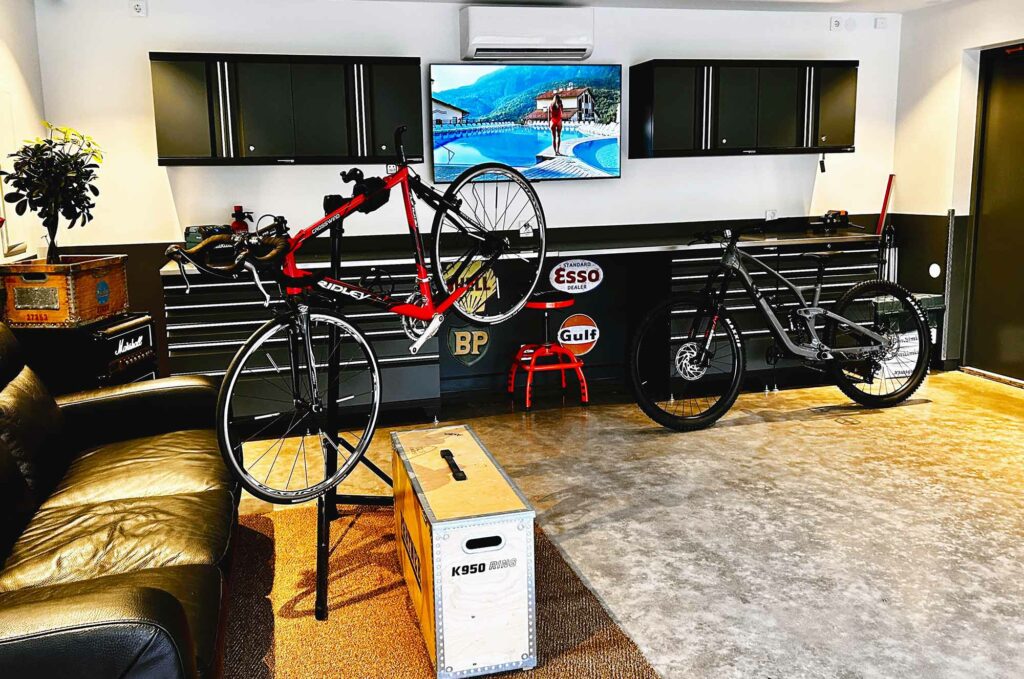 Ett modernt garage med två cyklar, en röd på stativ och en svart lutad mot ett skåp. En TV visar ett naturskönt landskap. Rummet har en svart soffa, förvaringsskåp, vintageskyltar och en liten trälåda.