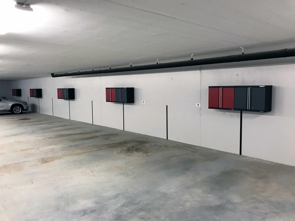 Ett rymligt, tomt underjordiskt garage har ett rent betonggolv och en elegant svart och röd väggskåp monterad längs den högra väggen. En silverfärgad bil är delvis synlig till vänster då takbelysning lyser upp området.