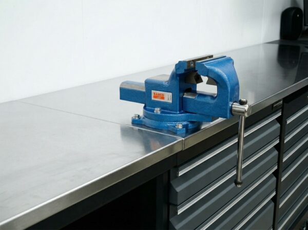 Ett skruvstäd i blå metall är monterat på en arbetsbänk i rostfritt stål med grå lådor undertill, i en ren och organiserad verkstadsmiljö.
