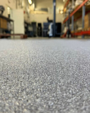 Närbild av ett strukturerat golv belagt med Epoxigolv PRO färgsand-paket i en lager- eller industrimiljö, med hyllor och utrustning suddade ut i bakgrunden.